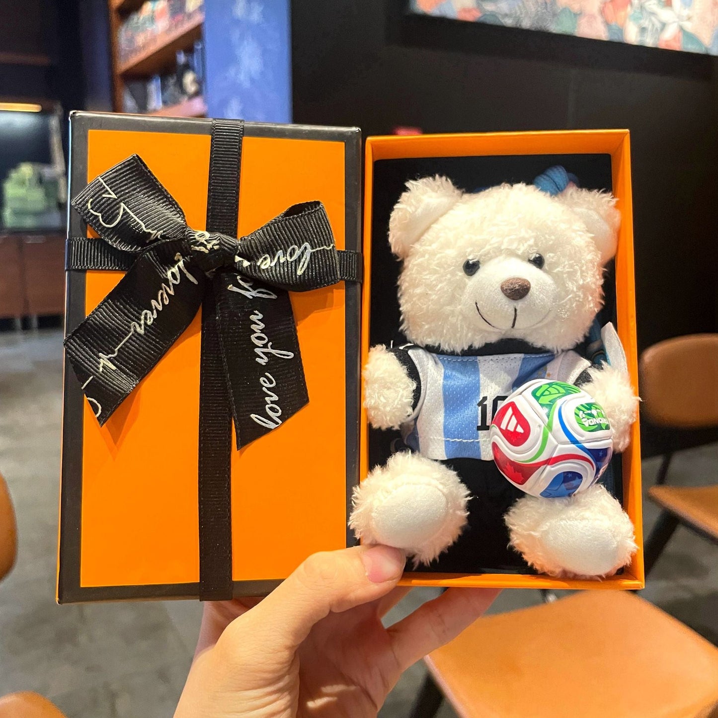 White plush bear keychain in orange gift box, blue jersey holding multicolor soccer ball