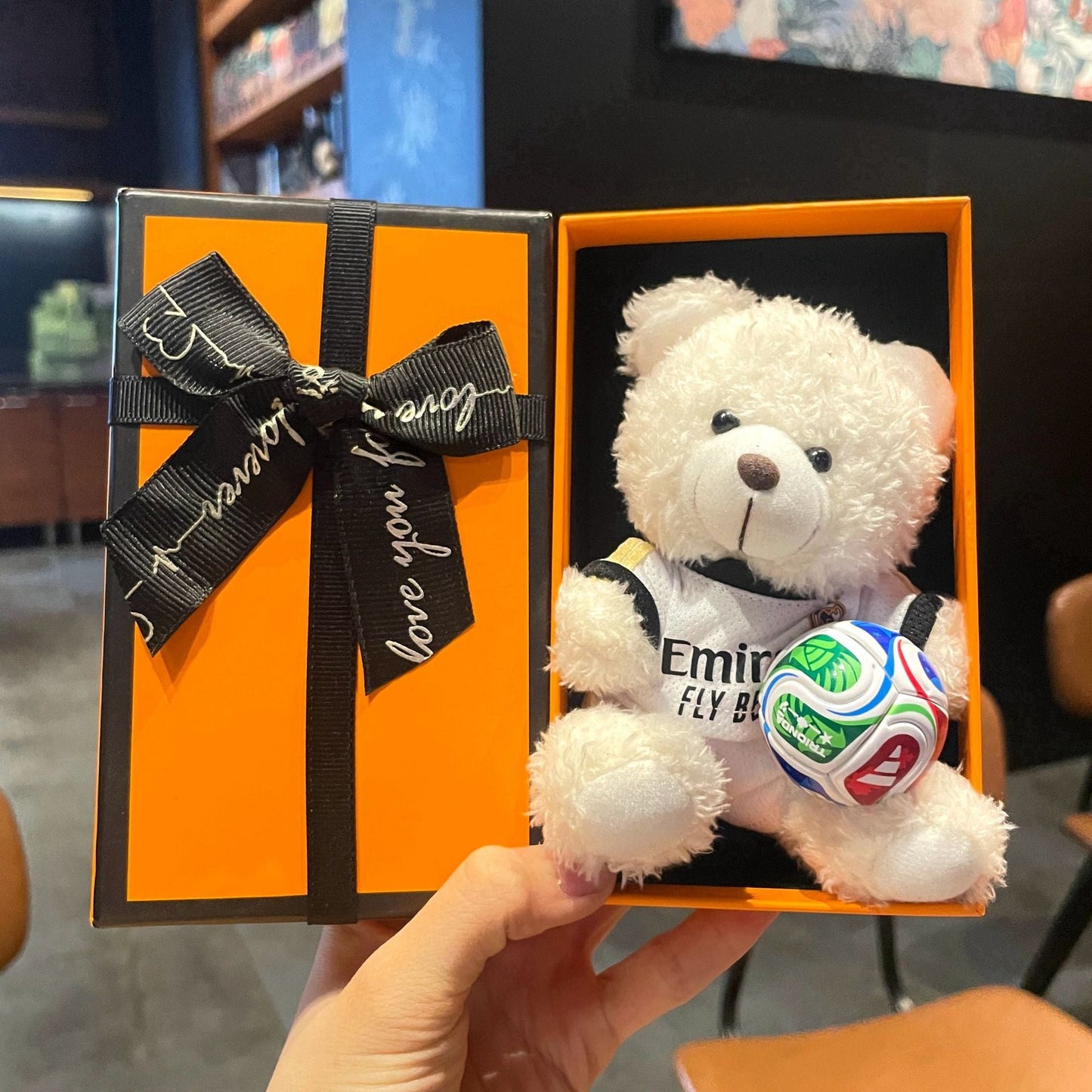 White plush bear keychain in orange gift box, “Emirates” jersey holding multicolor soccer ball