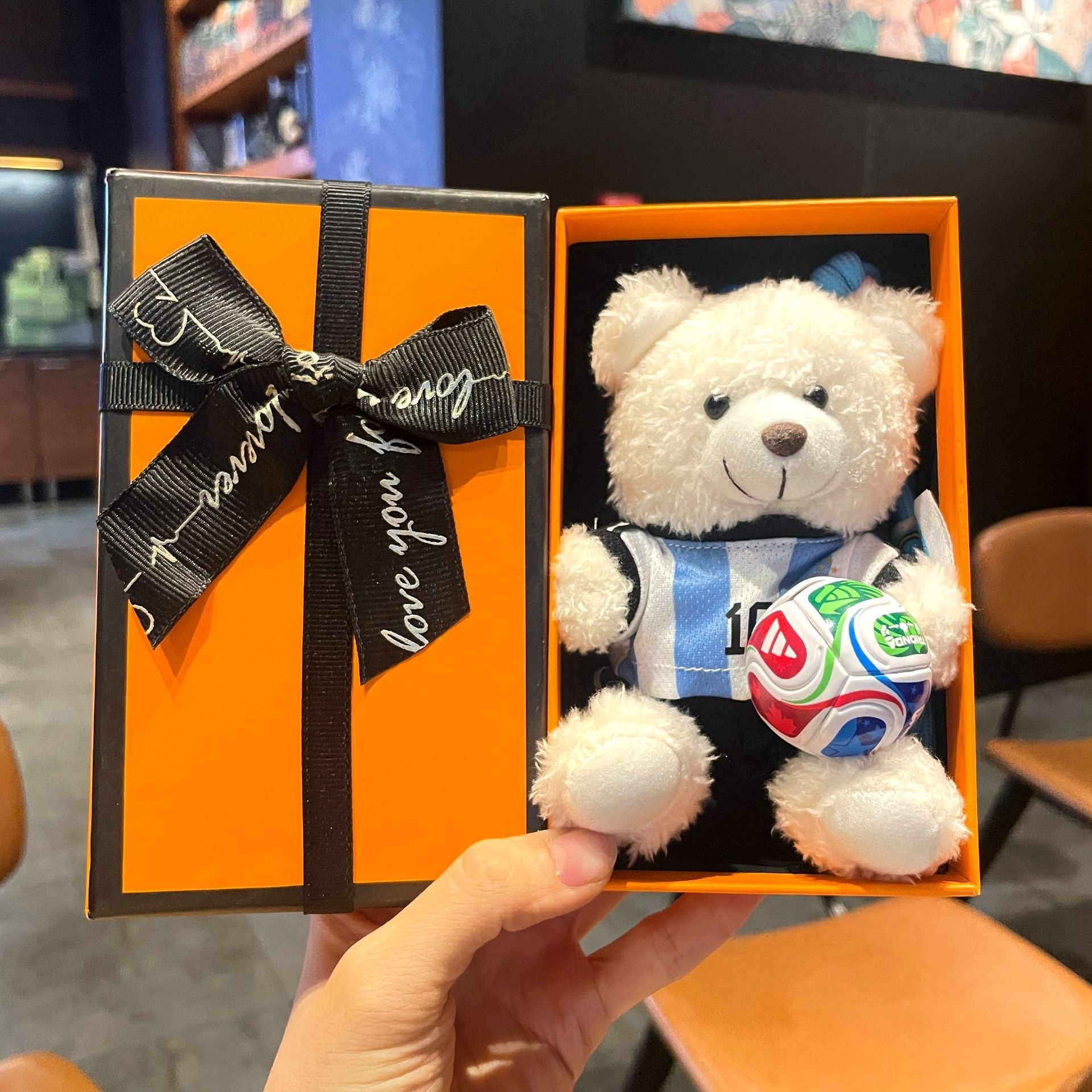 White plush bear keychain in orange gift box, blue jersey holding multicolor soccer ball