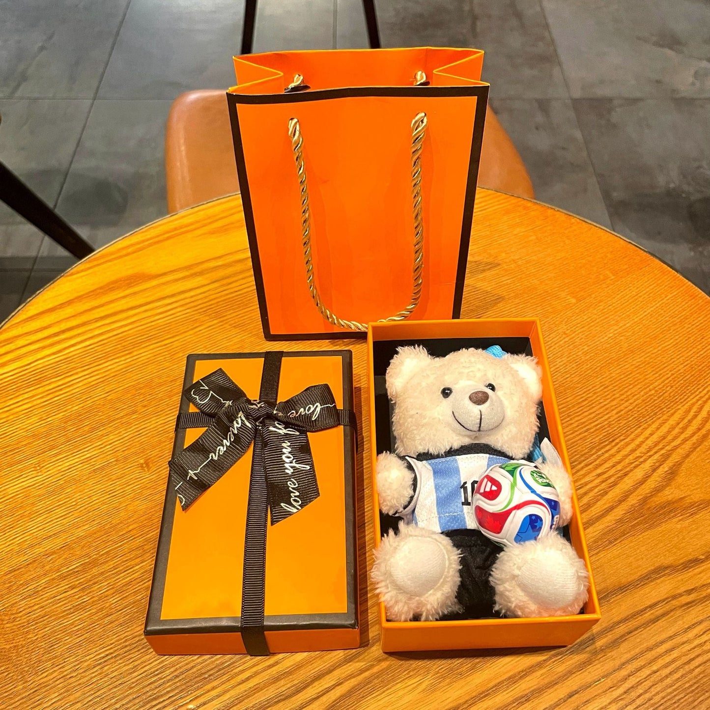White plush bear keychain in open orange gift box, wearing blue jersey with multicolor soccer ball