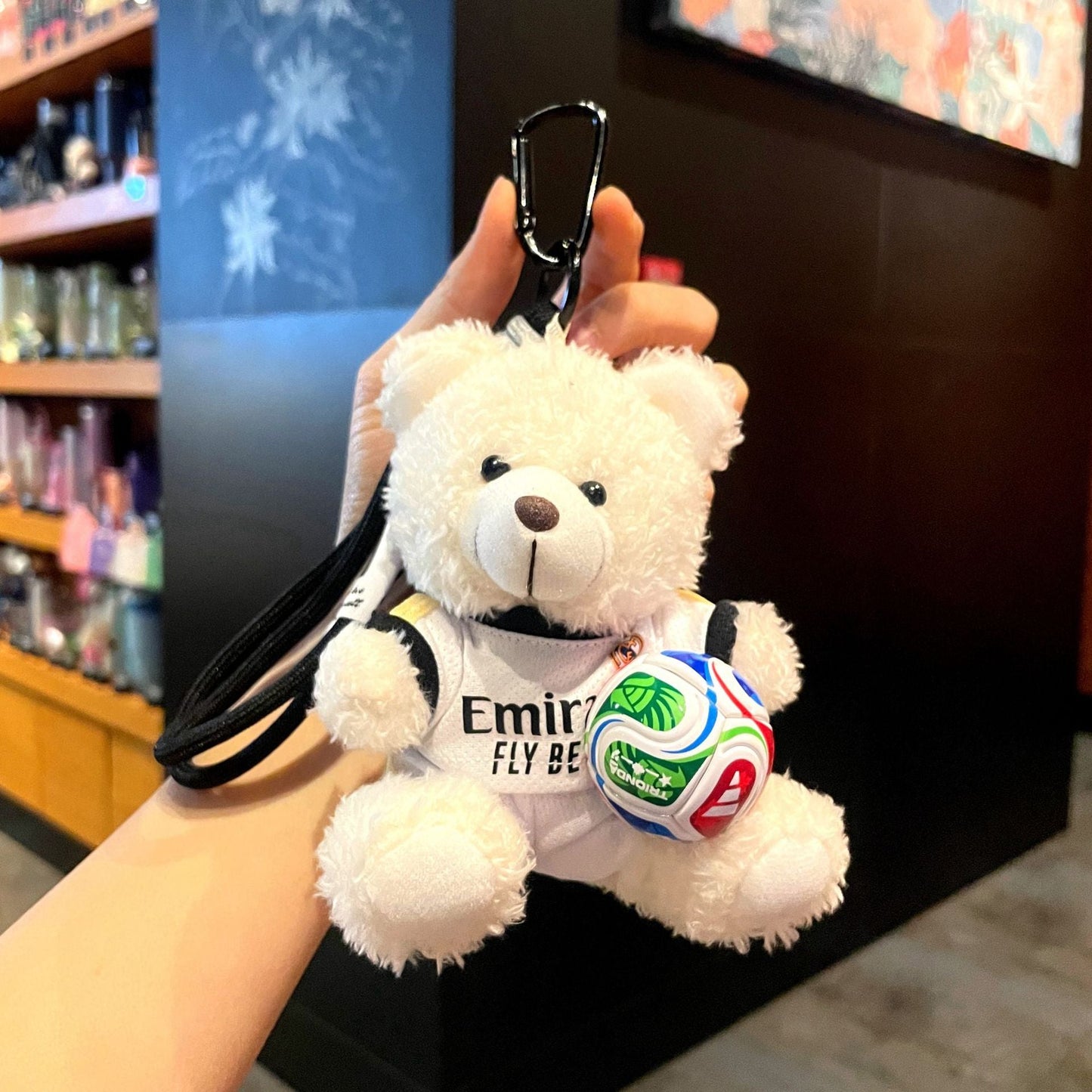 White plush bear keychain in white jersey with Emirates text, holding a soccer ball