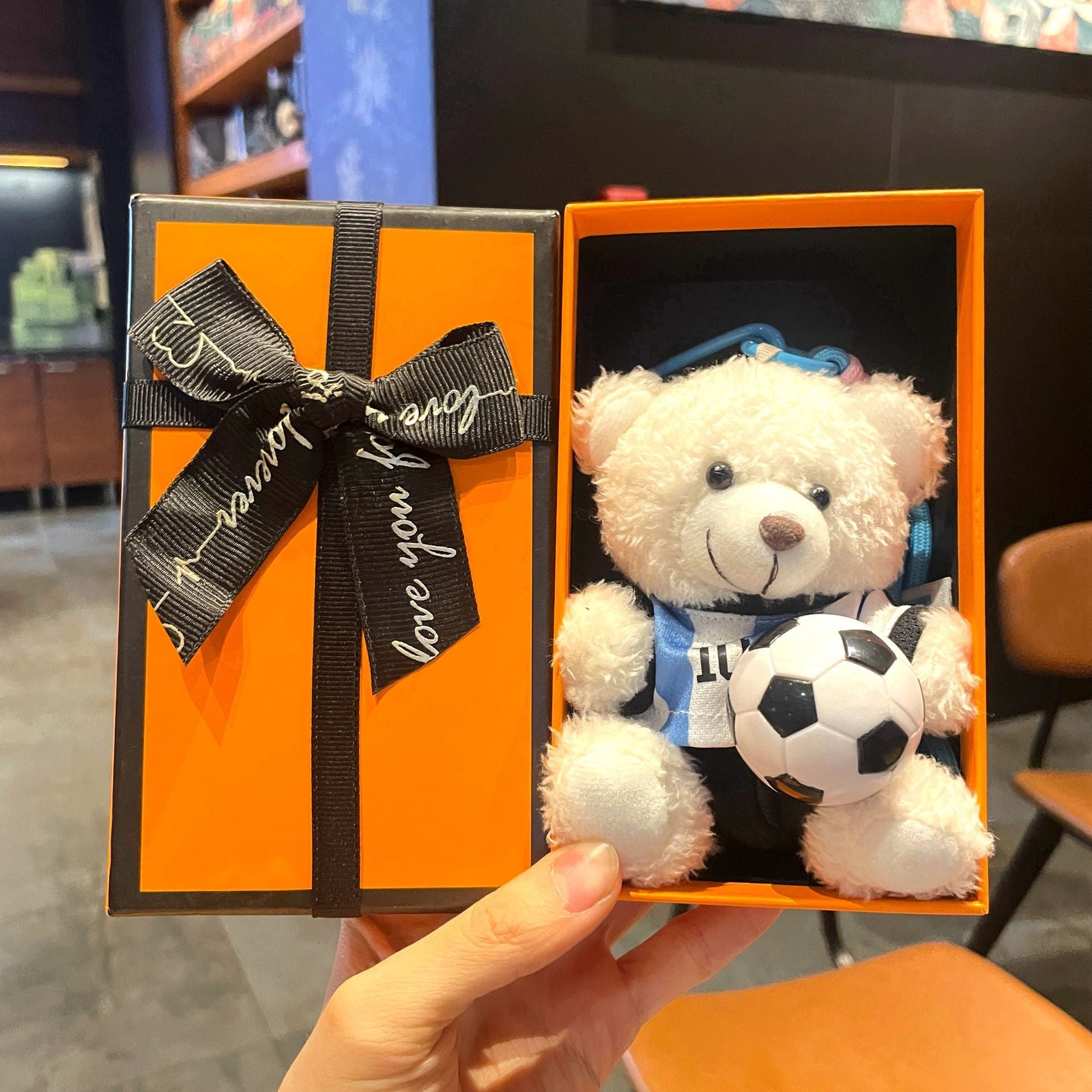 White plush bear keychain in orange gift box, wearing blue jersey and holding black-and-white soccer ball