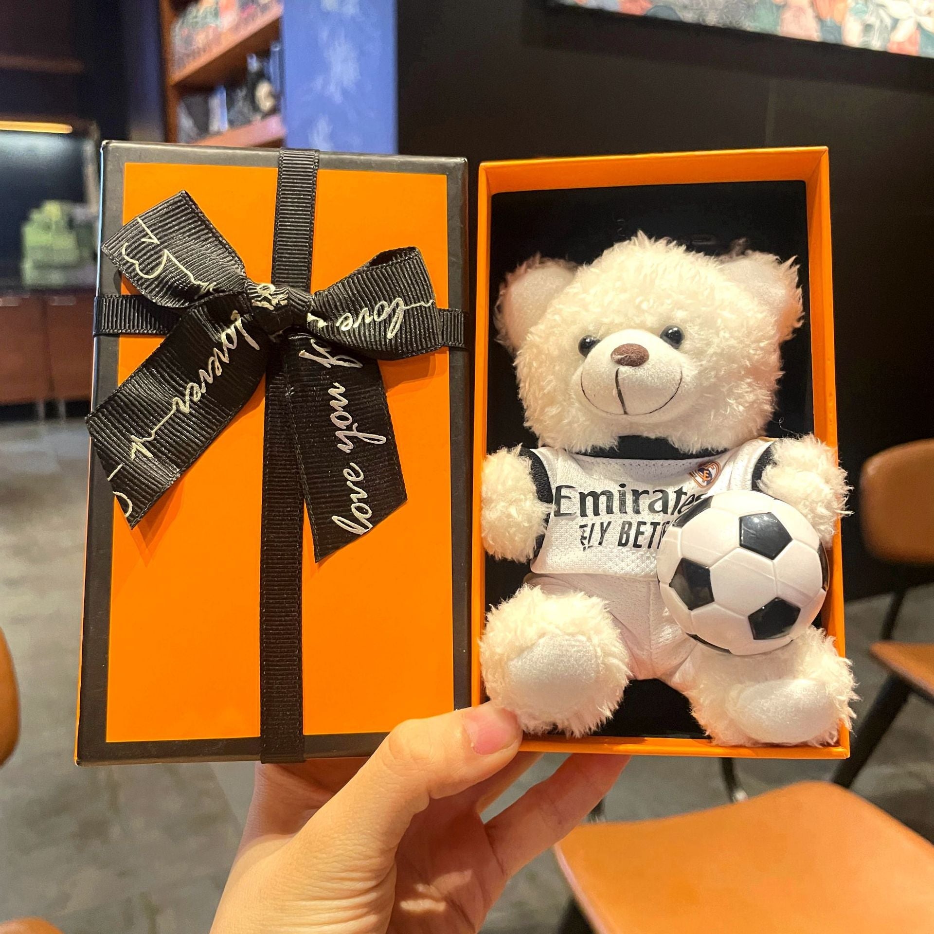 White plush bear keychain in orange gift box, “Emirates” jersey holding black-and-white soccer ball