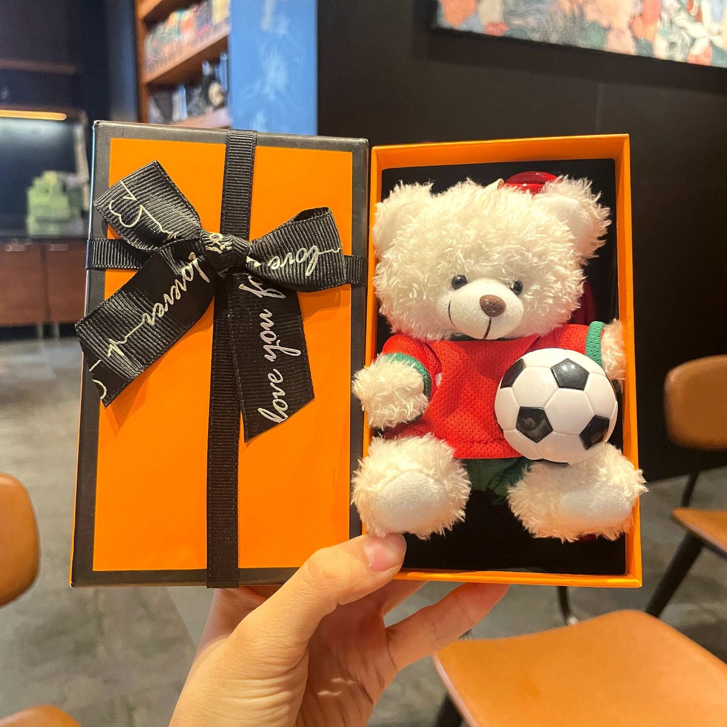 White plush bear keychain in orange gift box, red jersey holding black-and-white soccer ball