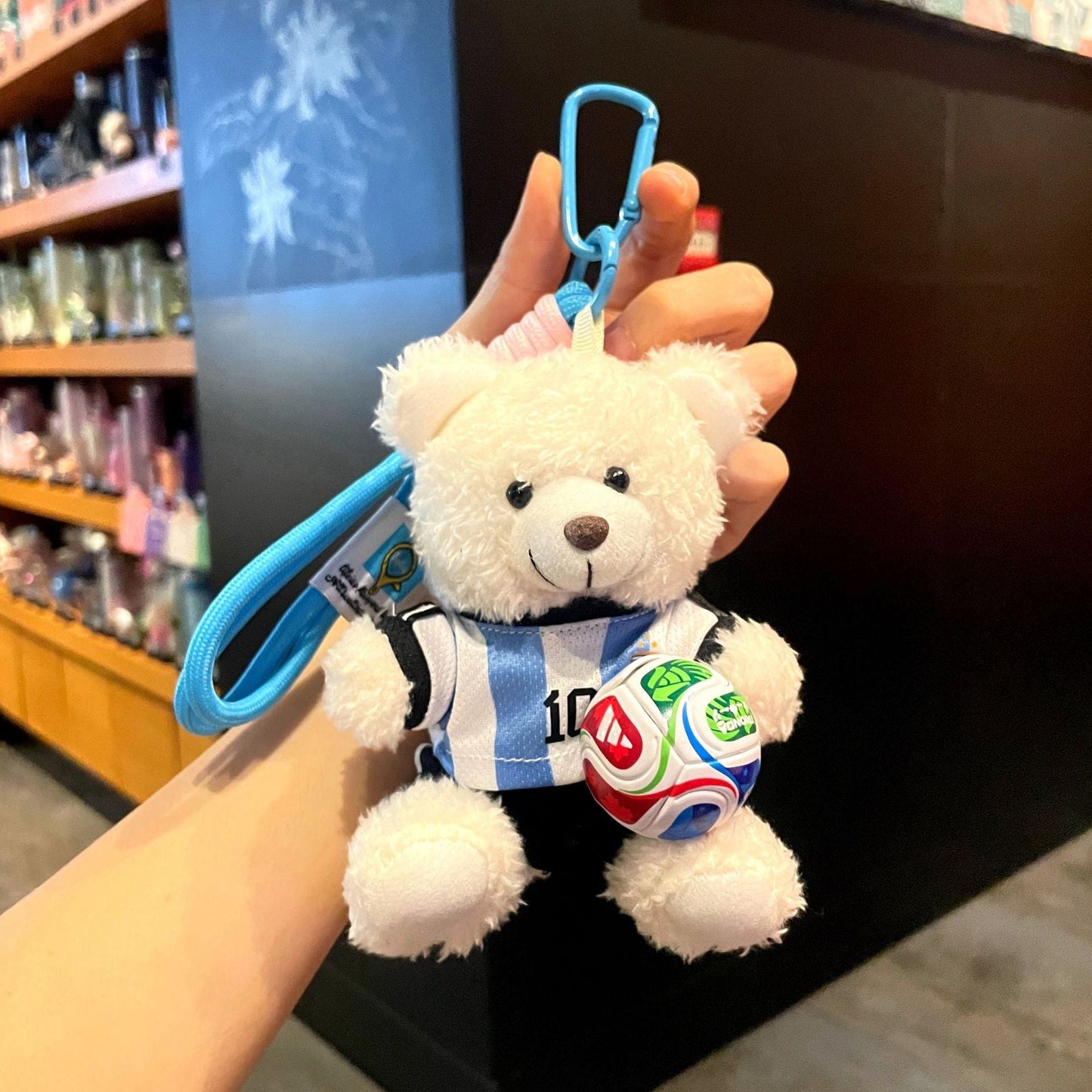 White plush bear keychain with blue strap, wearing blue soccer jersey, holding a red-and-blue ball