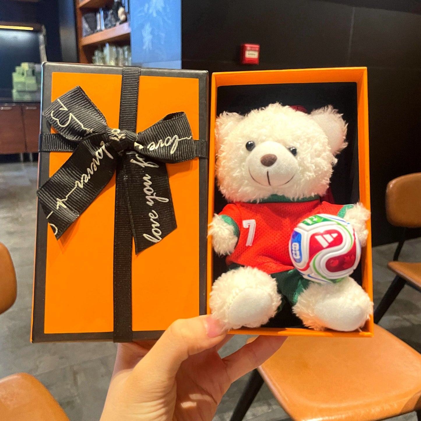 White plush bear keychain in orange gift box, red jersey holding multicolor soccer ball