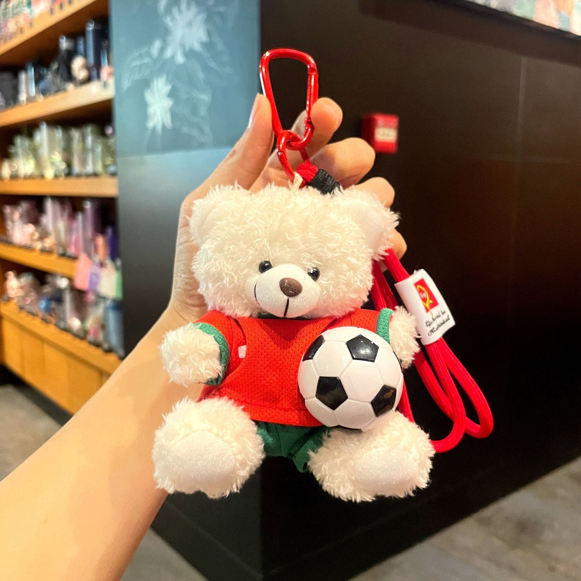 White plush bear keychain in red soccer jersey with green shorts, holding a black-and-white soccer ball