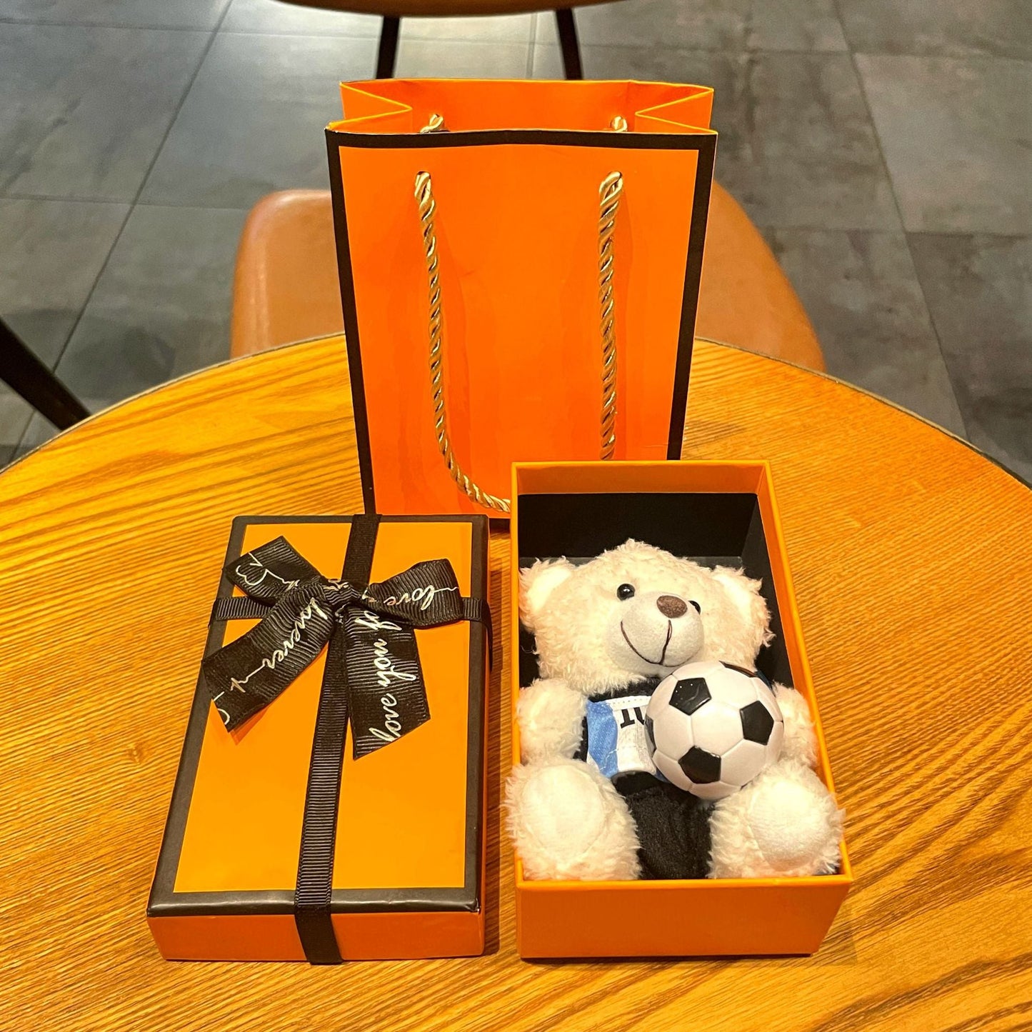 White plush bear keychain in open orange gift box, holding black-and-white soccer ball