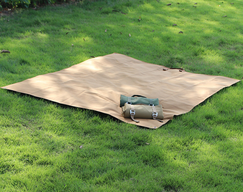 Tan waterproof picnic mat folded open on grass with attached carry bag and straps.